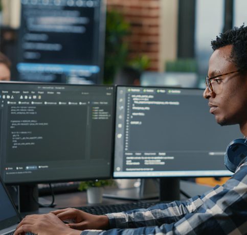 sofware-developer-thinking-while-touching-beard-while-typing-laptop-sitting-desk-with-multiple-screens-parsing-code-focused-database-admin-working-with-team-coding-background sofware-developer-thinking-while-touching-beard-while-typing-laptop-sitting-desk-with-multiple-screens-parsing-code-focused-database-admin-working-with-team-coding-background